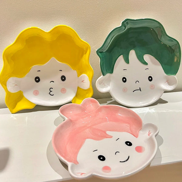 Adorable ceramic plates with cute face designs, featuring yellow, green, and pink hair, perfect for snacks or decor."