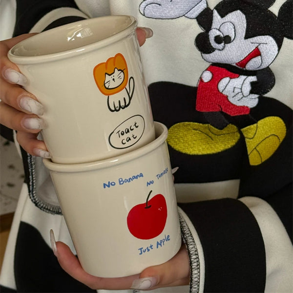 Adorable ceramic mugs with playful cartoon designs, including Toast Cat in a lion mane, an apple with "No Banana No Tomato" text, and colorful animal doodles.