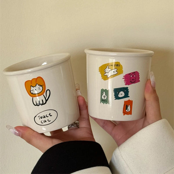 Adorable ceramic mugs with playful cartoon designs, including Toast Cat in a lion mane, an apple with "No Banana No Tomato" text, and colorful animal doodles.