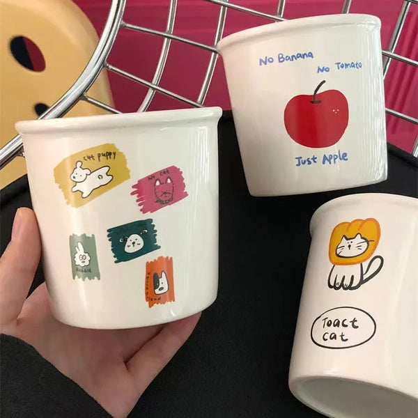 Three adorable ceramic mugs with playful cartoon designs, including Toast Cat in a lion mane, an apple with "No Banana No Tomato" text, and colorful animal doodles.